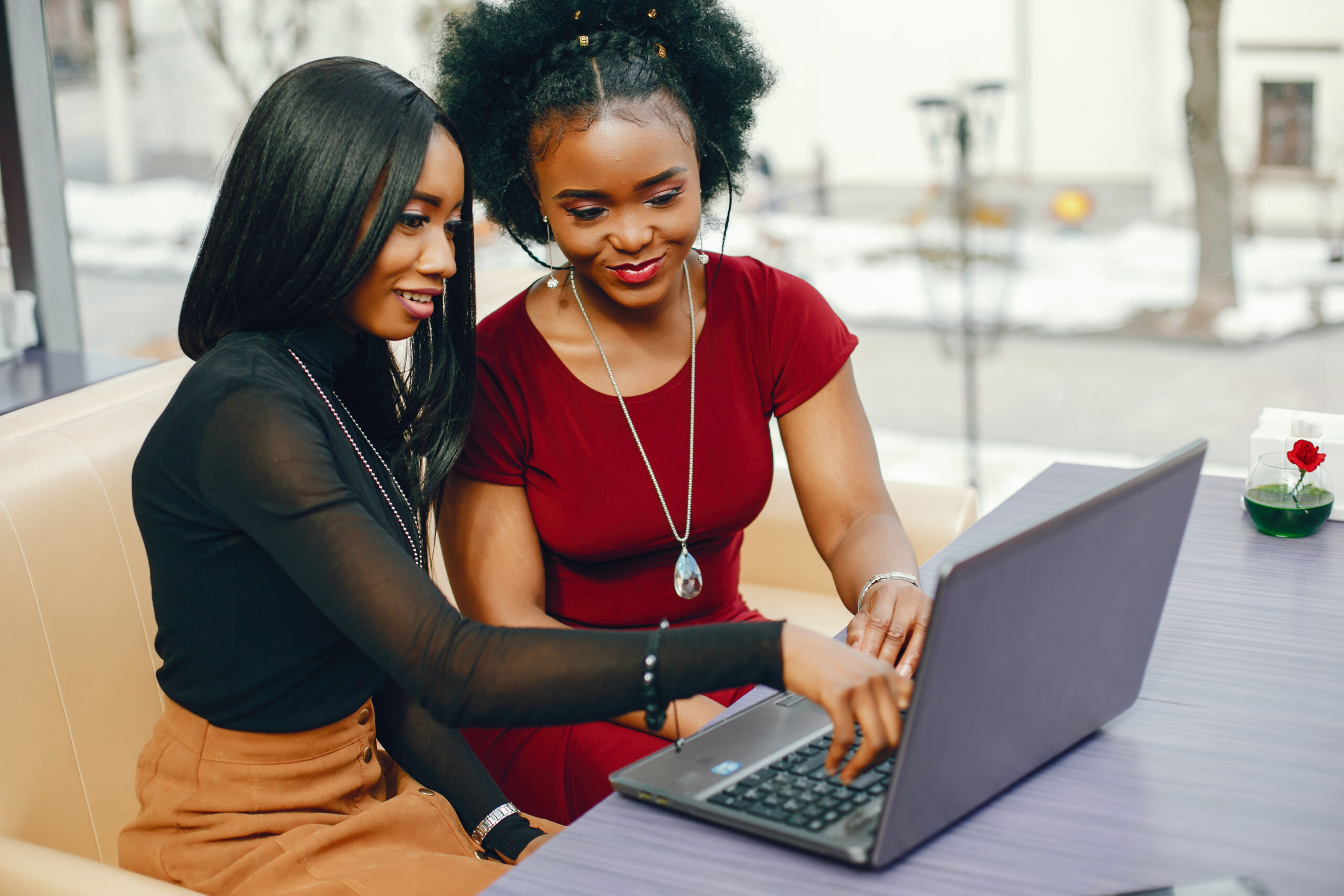 two beautiful and stylish young, dark girls sitting in a restaurant at the table, chatting and using a laptop